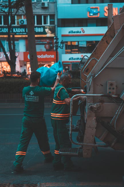Two waste collection workers wearing dark green uniforms with reflective orange stripes are handling blue plastic sacks filled with waste, which they are loading into a compact waste collection vehicl